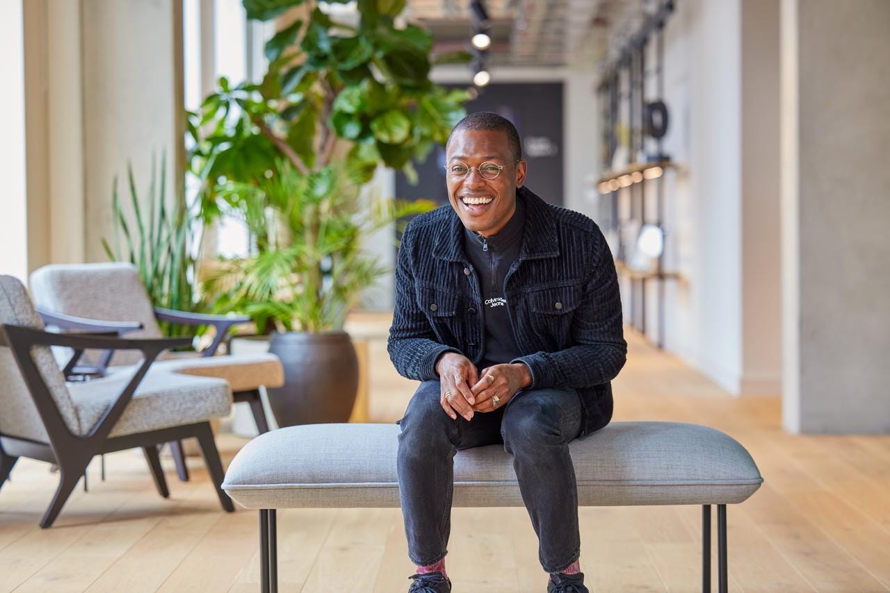 jai, smiling, sitting on a bench in a bright plant-filled studio
